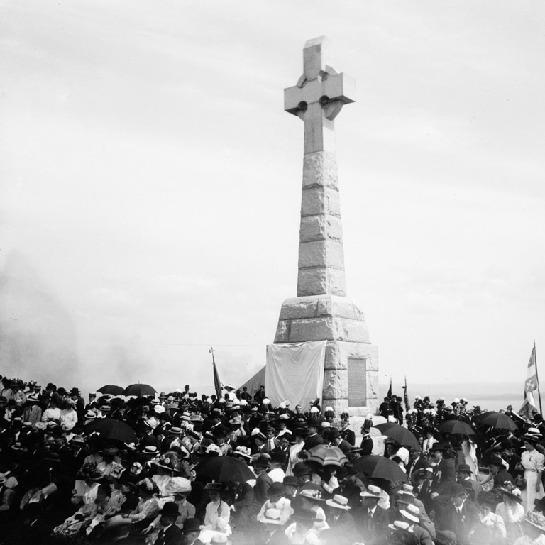 Photographie en noir et blanc d'une foule entourant un monument comportant une grande croix celtique.