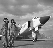 Photographie noir et blanc de deux hommes debouts à côté d'un avion regardant à l'horizon.