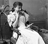 Photographie noir et blanc d’Émilie Lavergne portant une robe pâle, assise dans un fauteuil et lisant une lettre.