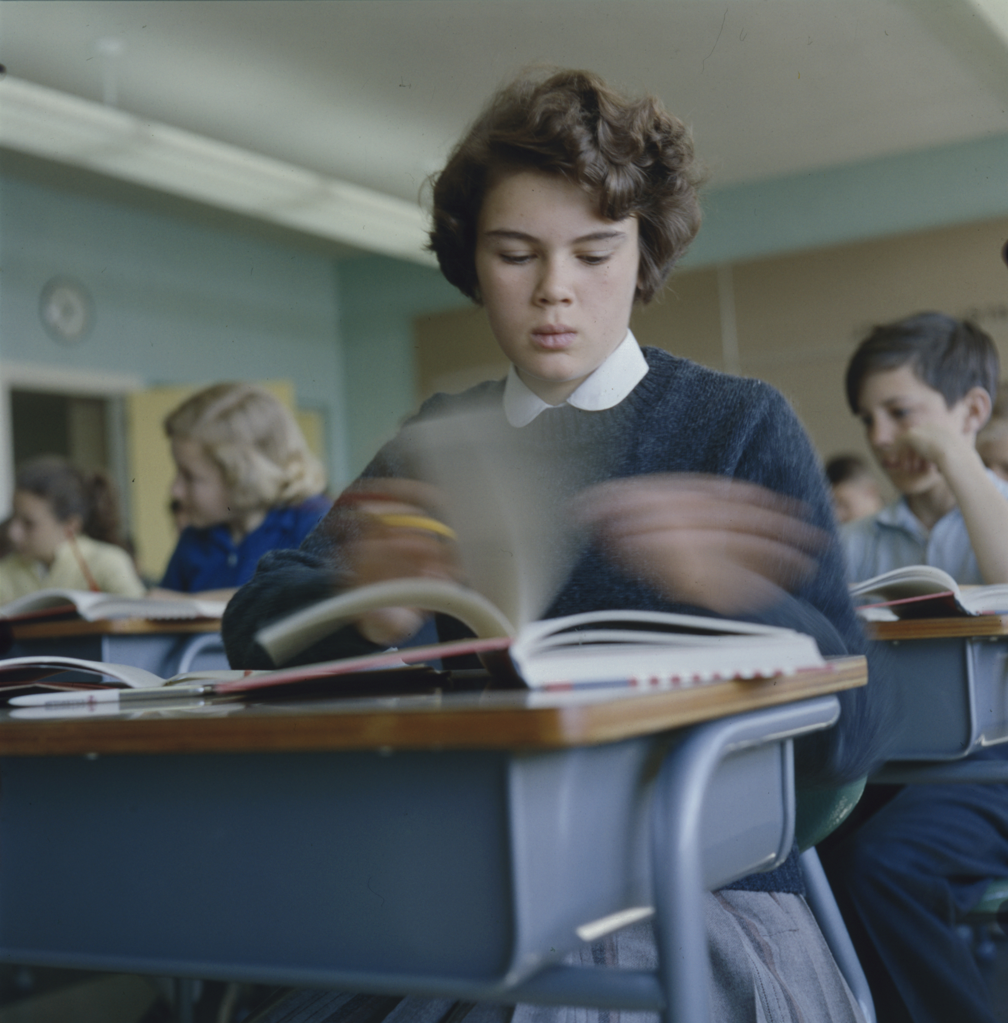 Jeune fille assise à un pupitre et tournant les pages d’un livre dans une salle de classe. On aperçoit d’autres enfants derrière elle.