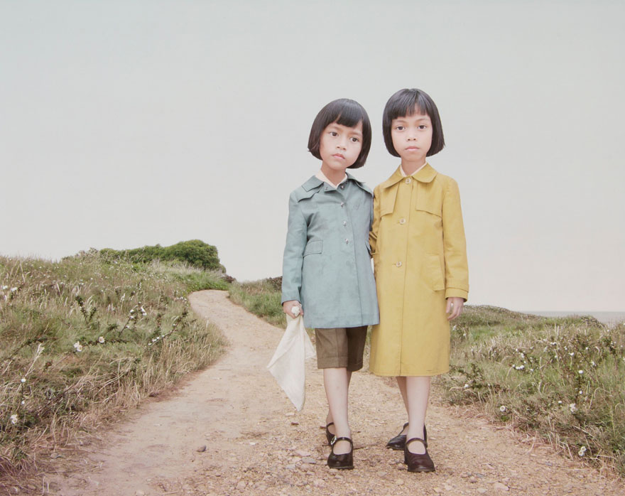 Deux petites filles debout sur un chemin de terre. L'une porte un manteau jaune, l'autre est vêtue d'un manteau bleu et tient un linge blanc.