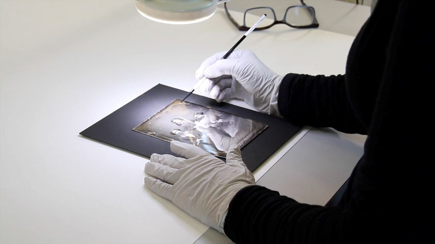 On utilise un pinceau sous microscope pour enlever les moisissures sur une photographie montrant trois jeunes filles (deux assises, l'autre debout au centre).