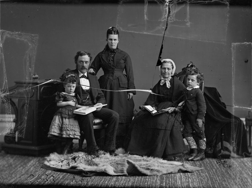 Image positive d'un négatif sur plaque de verre. La photo est un portrait de famille. Une petite fille debout lit un livre avec son père, qui est assis. À côté de lui, son épouse est debout. Une dame âgée est assise à côté d'elle, avec un petit garçon debout à ses côtés.