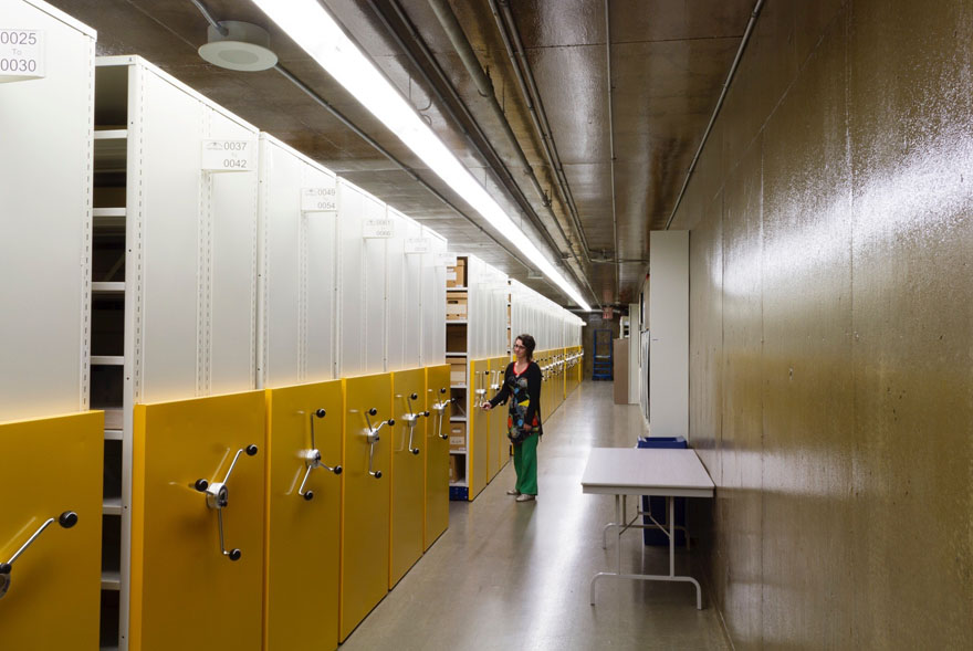 Longue chambre forte équipée de rayonnages mobiles; une femme déplace un rayonnage.