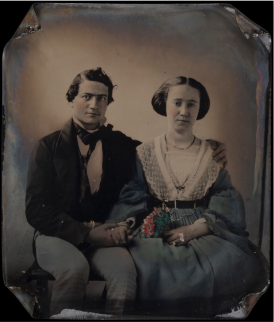 Portrait d'un jeune couple. L'homme entoure les épaules de la femme d'un bras et tient sa main de l'autre. La jeune femme tient un bouquet de fleurs. Les quatre coins de la plaque photographique sont pliés vers l'intérieur.