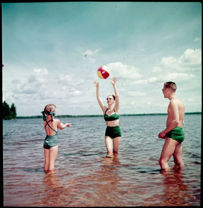 Une petite fille, une femme et un homme, tous en maillot de bain, se lancent un ballon de plage en eau peu profonde.