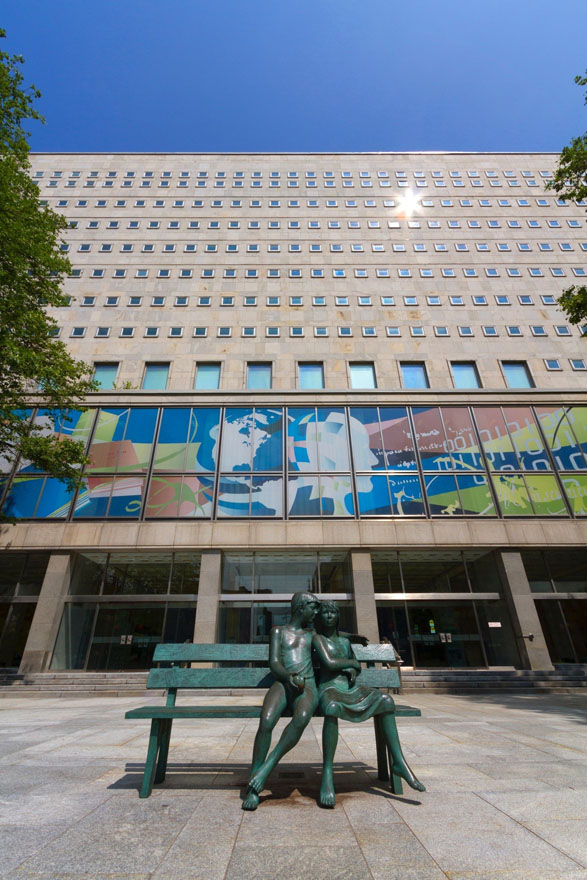 Façade d'un grand édifice avec, à l'avant-plan, une statue de bronze représentant deux personnes assises sur un banc.