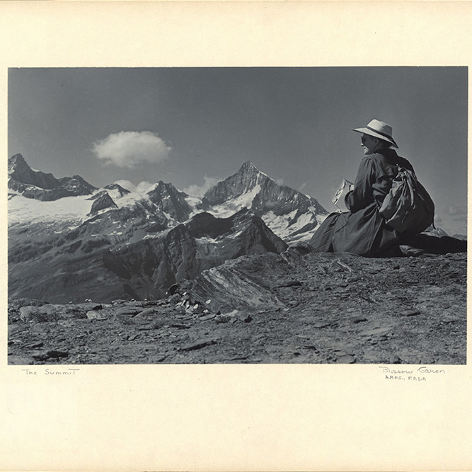 Photo noir et blanc montrant une dame assise devant un panorama de montagnes rocheuses partiellement enneigées.